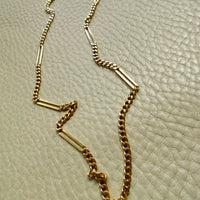 An 18k gold chain necklace with alternating small curb links and elongated bar links, laid out on a textured off-white surface.