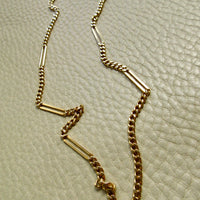 An 18k gold chain necklace with alternating small curb links and elongated bar links, laid out on a textured off-white surface.