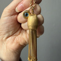Gold tassel necklace with an eye design held by a hand against a gray background