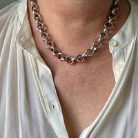 A close-up of a person wearing a chunky silver chain necklace with large interlocking links, styled over a white button-down shirt.