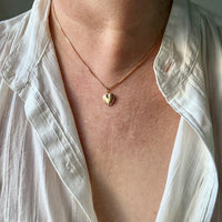 Person wearing a gold necklace with a heart pendant, visible under a white shirt.