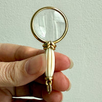 Hand holding a small magnifying glass with a decorative handle against a light background