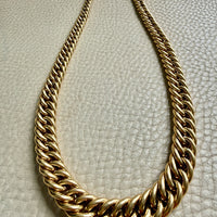 A thick, chunky 18k gold curb link chain necklace displayed on a textured off-white leather surface.