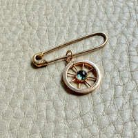 Gold safety pin with a wheel charm and blue gemstone on a textured beige background