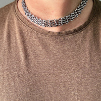 A close-up of a person's neck and upper chest wearing a chunky vintage silver chain choker with interlocking chain links, paired with a brown knit t-shirt.