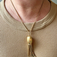 Gold necklace with a tassel pendant worn by a person in a beige shirt.