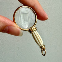 Hand holding a small magnifying glass with a gold frame and white handle against a light background