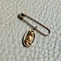 Gold safety pin with a gold owl pendant on a textured gray surface