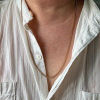 A close-up of a person's neck and chest wearing a delicate gold chain necklace over an open white linen button-up shirt.