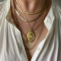 A person wearing multiple gold necklaces with a prominent gold medallion pendant in the center, featuring blue enamel detail.