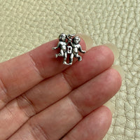 A small silver charm depicting two gemini figures, held between a person's fingers against a textured beige background.