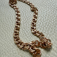 A chunky rose gold chain necklace displayed on a textured off-white surface.