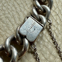 Close-up of a silver chain bracelet clasp with engraving for Minnie, on a beige leather surface