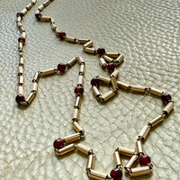 An 18k gold chain necklace with cylindrical links and small dark red gemstone accents, laid out on a textured light beige surface.