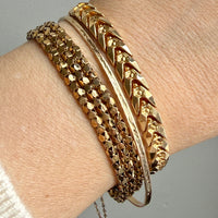 A close-up of a person's wrist wearing a stack of vintage 18k gold bracelets, including a faceted chain bracelet, a textured bangle, a smooth bangle, and a chevron-patterned bangle.