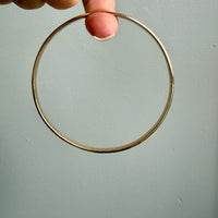 A person's hand holding a narrow, polished gold bangle against a plain, light gray wall.