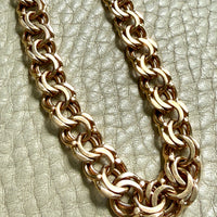 A close-up of a chunky 18k gold chain link necklace resting on a textured light beige leather surface.