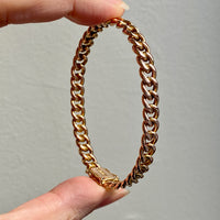 A vintage rigid curb link hinged bangle bracelet in rosy 14k gold, held between fingers.