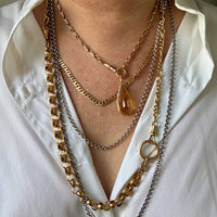 A close-up of a person wearing a layered necklace stack featuring multiple gold and silver-tone chains, including a teardrop pendant, on a white collared shirt.