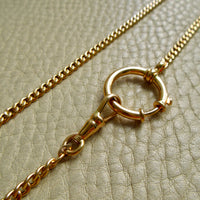 A close-up of an 18k gold chain necklace with a circular bolt ring clasp, laid on a textured beige leather surface.