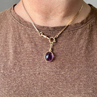 Gold necklace with a purple gemstone pendant worn by a person in a brown shirt.