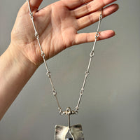 Hand holding a silver necklace with a unique pendant against a neutral background