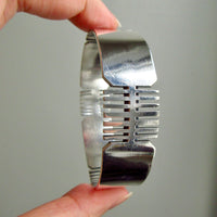 A hand holding a polished silver bangle bracelet with a geometric, ribbed design against a plain gray background.