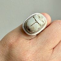 A close-up of a hand wearing a chunky silver ring with a textured, cracked stone scarab beelte inlay.