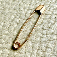 An 18k solid gold safety pin brooch resting on a textured light beige leather surface.