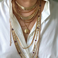 A close-up of a person wearing a layered stack of gold-toned necklaces over a white button-up shirt. The necklaces include chain styles, mesh details, beads, and a round locket pendant.