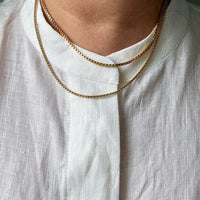 A close-up of a person wearing two gold chain necklaces layered over a white linen shirt.