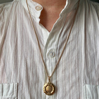 A close-up of a person wearing a gold pendant necklace with a textured circular medallion over a white button-up shirt.