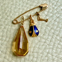A gold safety pin brooch with three dangling charms: a large faceted amber-yellow teardrop crystal, a small gold and blue enamel ladybug charm, and a small gold cava glass charm, displayed on a textured off-white leather surface.