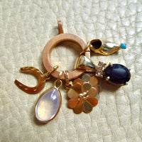 A gold charm cluster featuring a large circular ring, a horseshoe, a flower-shaped charm, a teardrop moonstone pendant, a small bird charm, and a dark oval sapphire gemstone, displayed on a textured beige leather background.