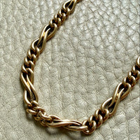 A close-up of an antique 18k gold watch chain necklace with a twisted, rope-like link design, resting on a textured light beige leather surface.
