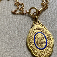 An 18k gold vintage medallion pendant with blue enamel detail, featuring floral wreaths and an ornate central design, mounted on a gold chain. penelopepenelope.com