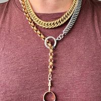 A close-up of a person wearing a layered necklace set featuring a thick gold Cuban link, a silver-toned chain, and a mixed gold and silver Y-necklace with a dangling ring charm.