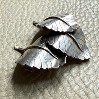 A sterling silver metal brooch shaped like overlapping leaves, photographed on a textured light beige leather surface.