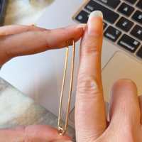 Gold safety pin held between fingers with a laptop in the background