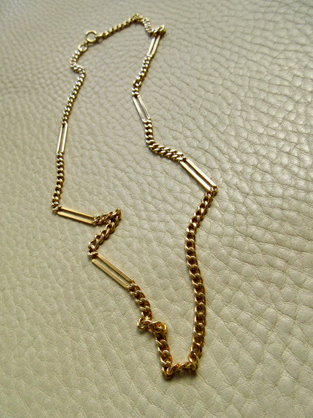 An 18k gold chain necklace with alternating small curb links and elongated bar links, laid out on a textured off-white surface.