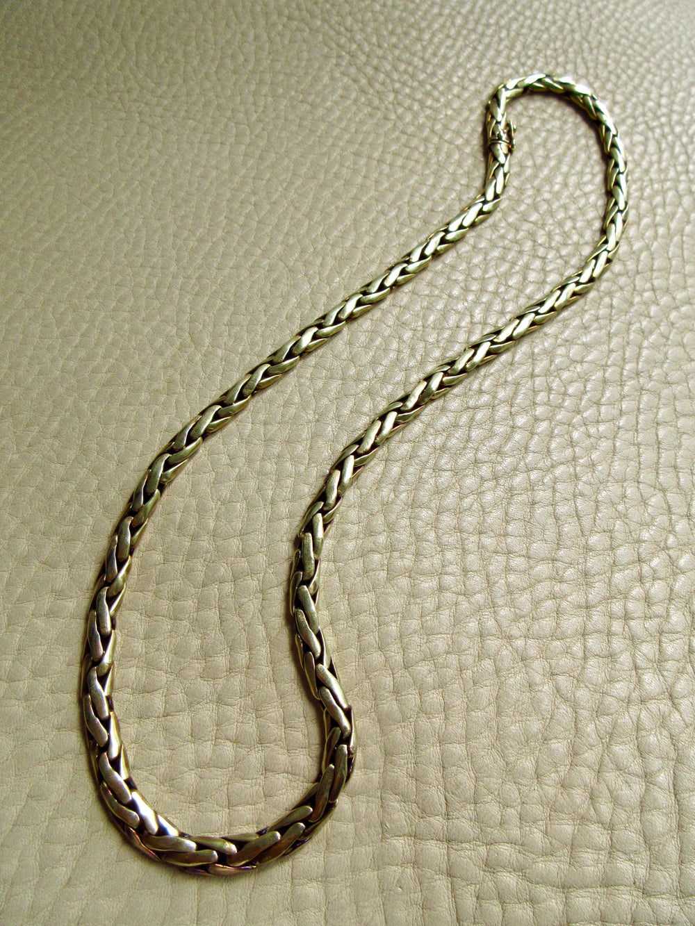 A braided 14k gold chain necklace with laid out on a textured beige leather surface.