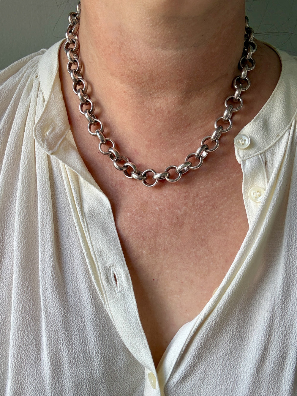 A close-up of a person wearing a chunky silver chain necklace with large interlocking links, styled over a white button-down shirt.