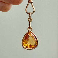 Gold pendant with a teardrop-shaped yellow gemstone held by a hand against a neutral background