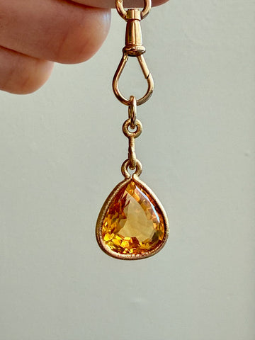 Gold pendant with a teardrop-shaped yellow gemstone held by a hand against a neutral background
