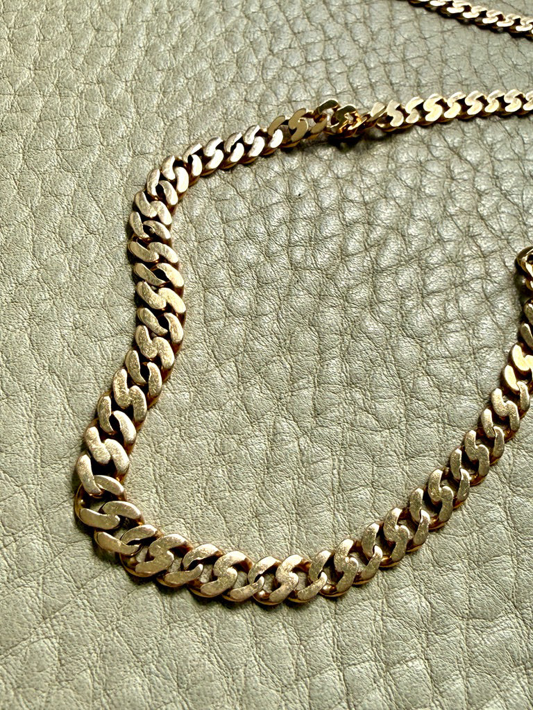 An 18k gold pressed curb link chain necklace draped across a textured light beige leather surface.