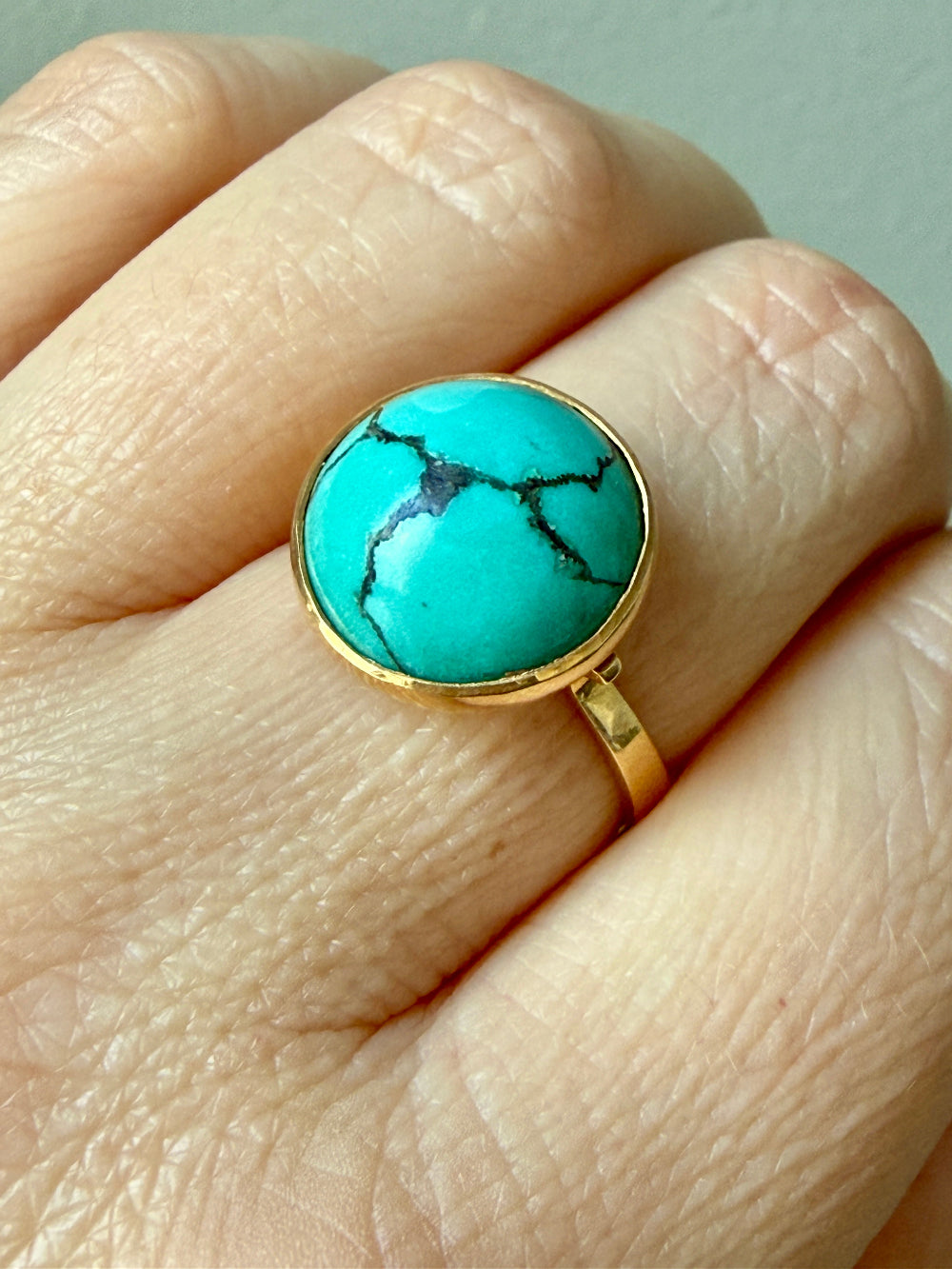 A gold ring with a round turquoise cabochon stone featuring black matrix veining, shown worn on a person's finger.