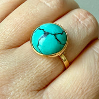 A gold ring with a round turquoise cabochon stone featuring black matrix veining, shown worn on a person's finger.