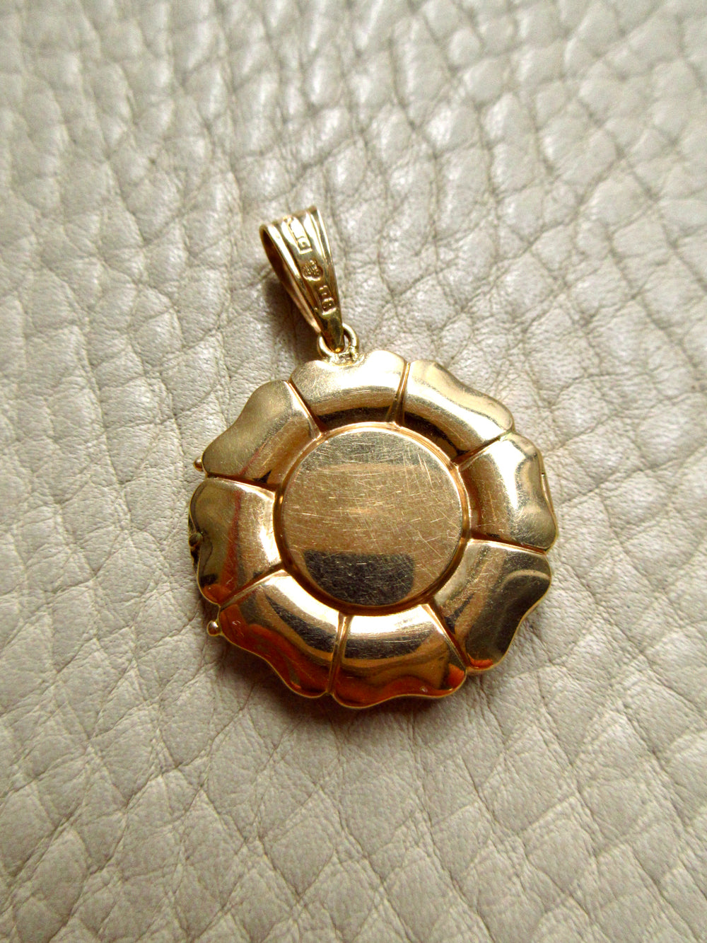 An 18k gold pendant with a circular sunburst or flower-like design and a smooth central disc, photographed on a textured off-white surface.