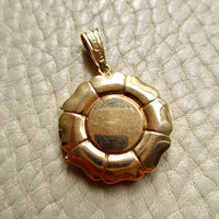 An 18k gold pendant with a circular sunburst or flower-like design and a smooth central disc, photographed on a textured off-white surface.