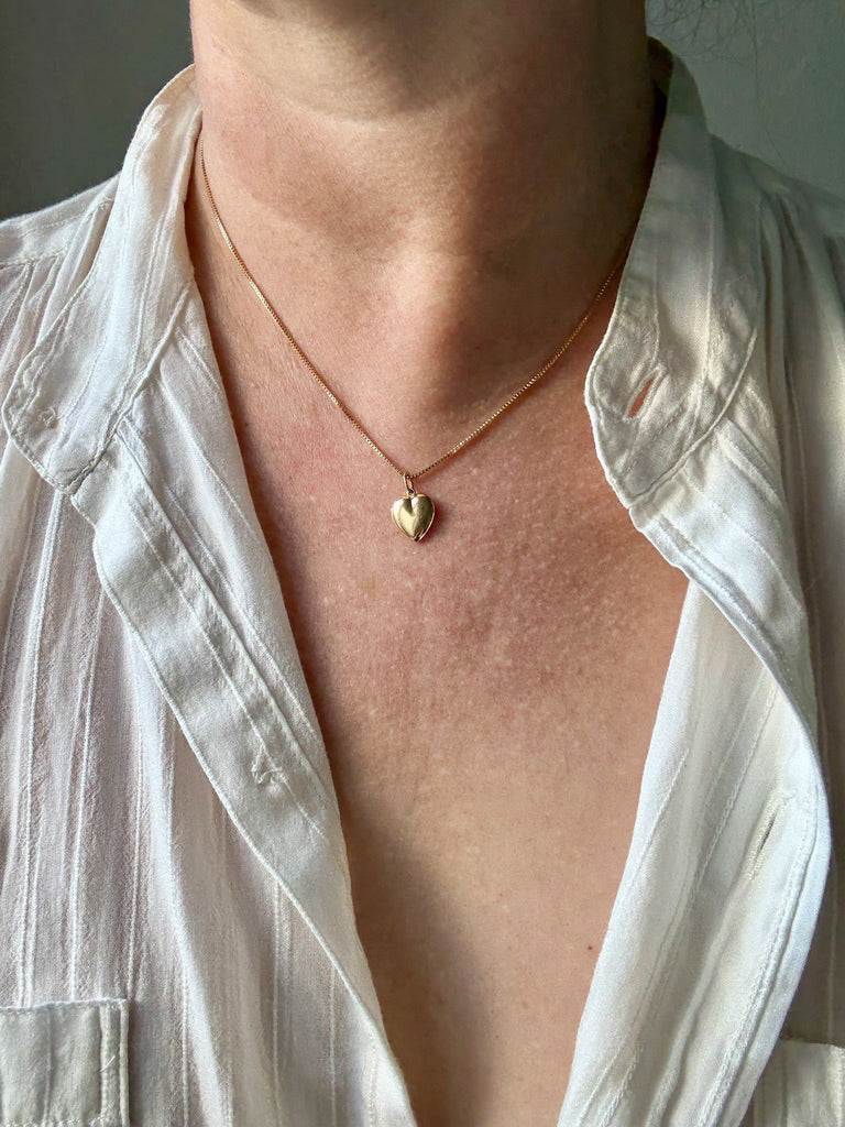 Person wearing a gold necklace with a heart pendant, visible under a white shirt.